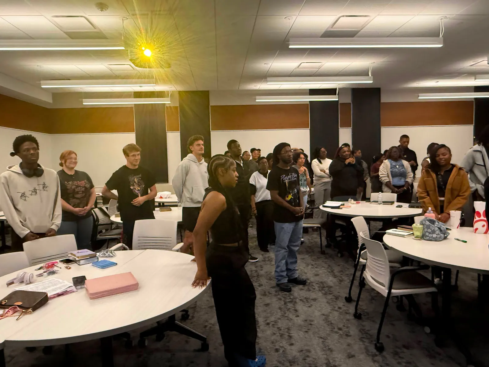 Students standing around round tables in a wide room, talking in groups before a session starts.