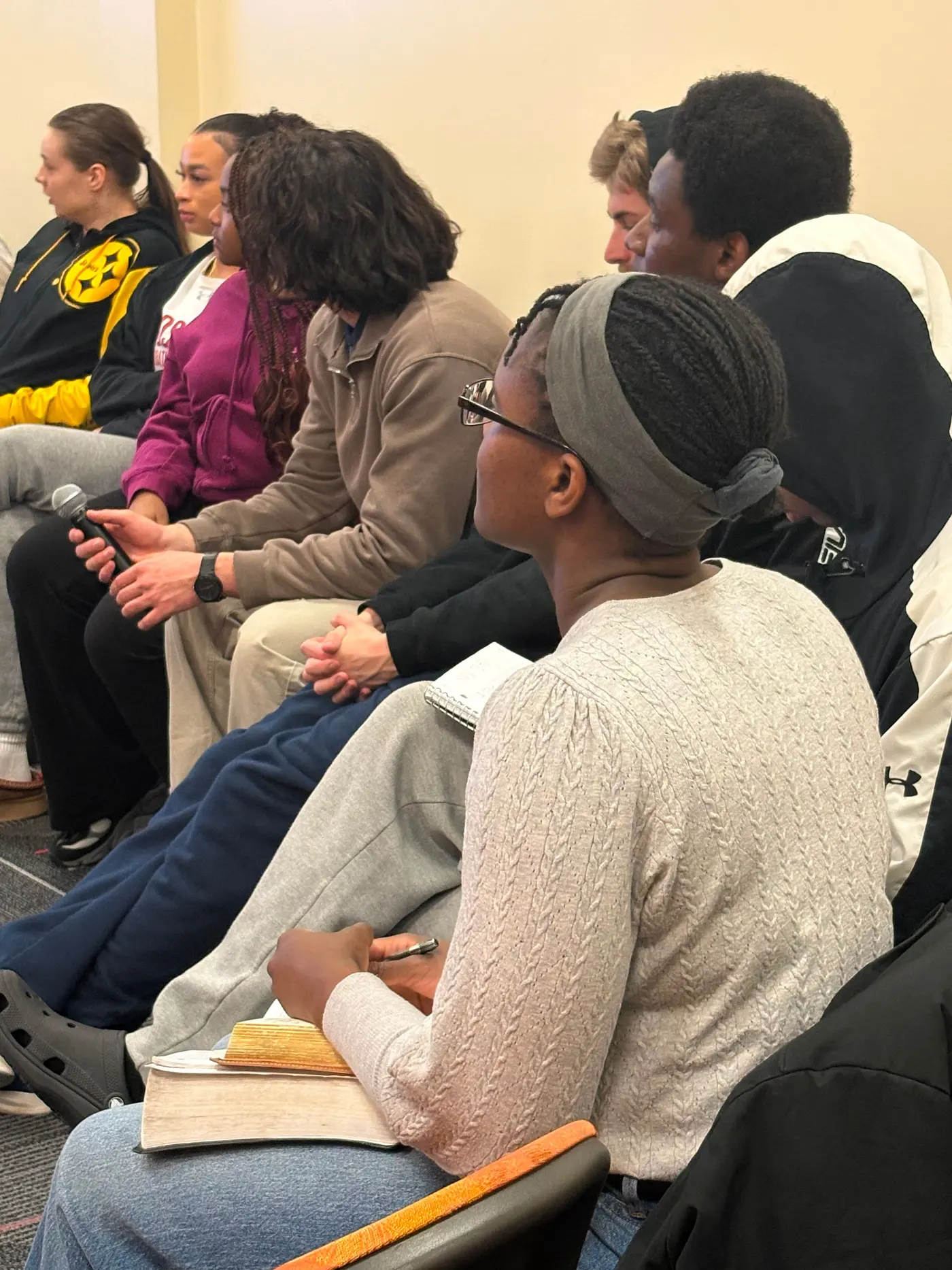 Students seated close together in a row, one of them holding an open book on their lap.