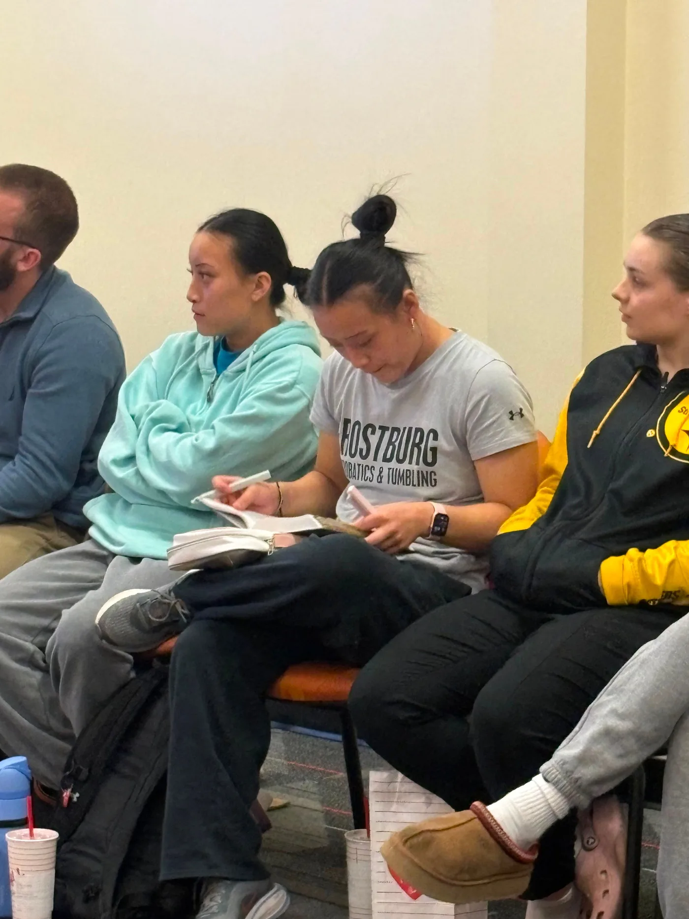 Four students seated along a wall, two of them writing in notebooks resting on their knees.