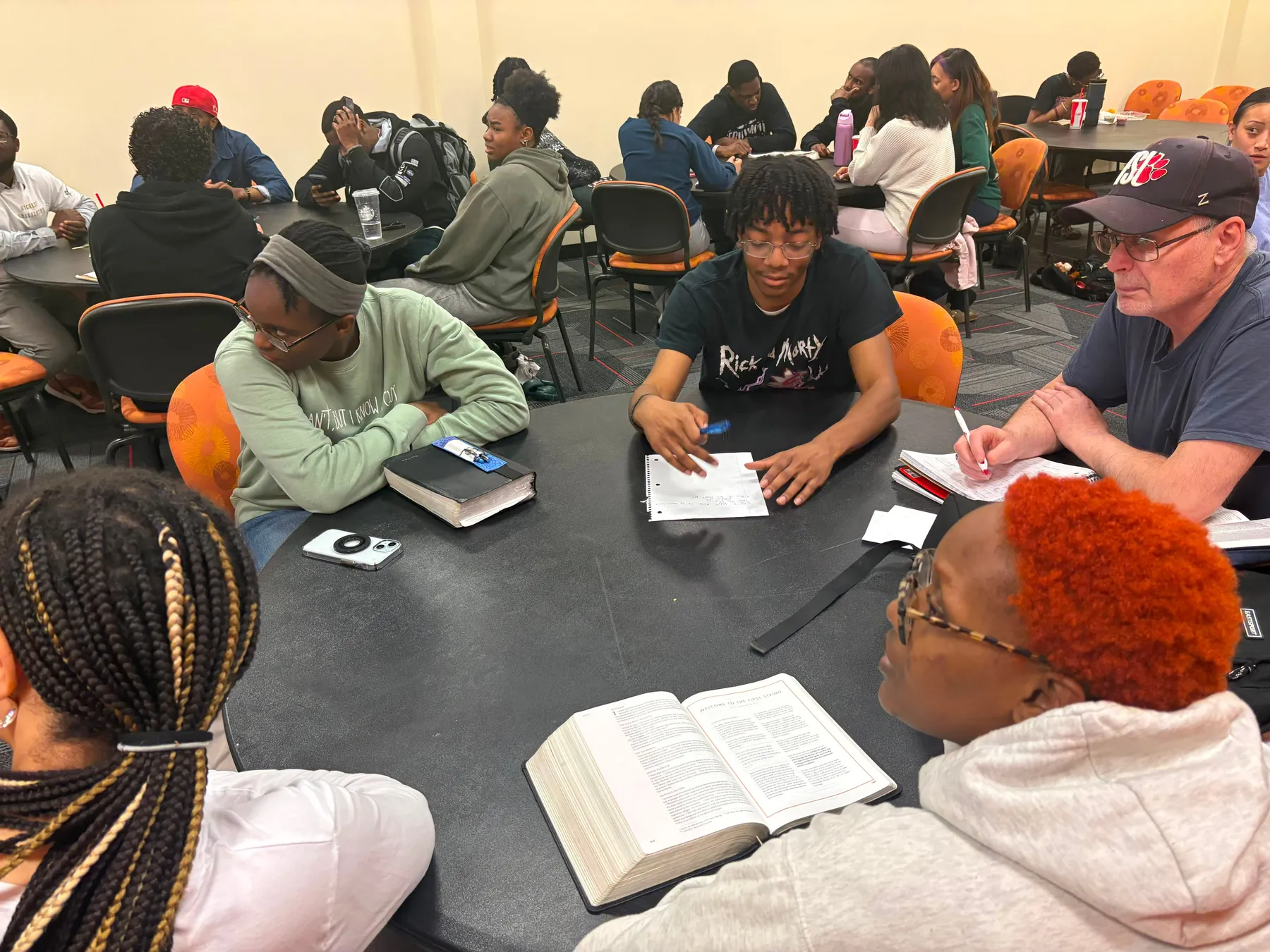 Students at round tables with open Bibles, notebooks and study guides in front of them, one Bible open in the foreground.