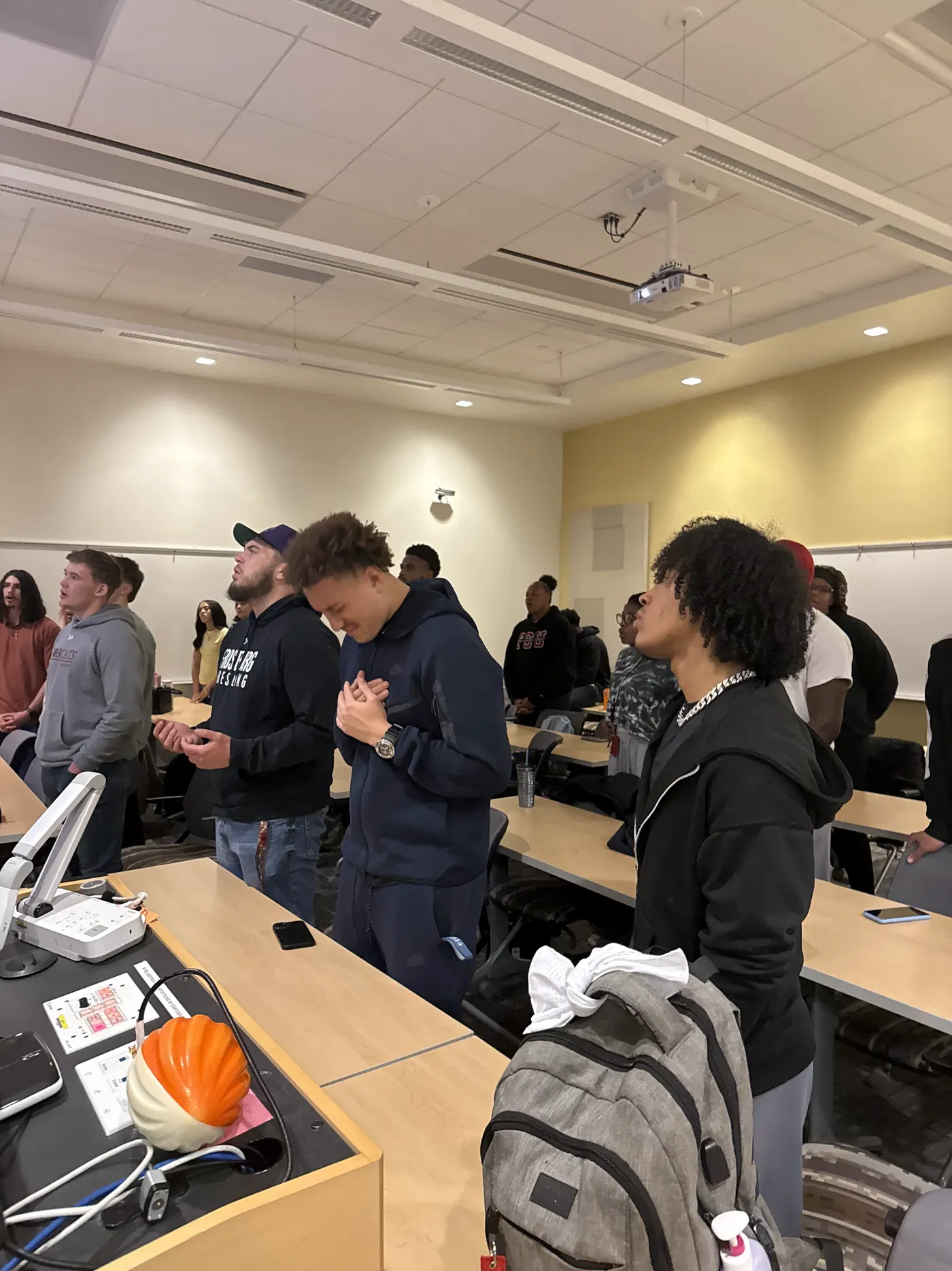 Students standing in a classroom with their hands clasped in front of them and their eyes closed.