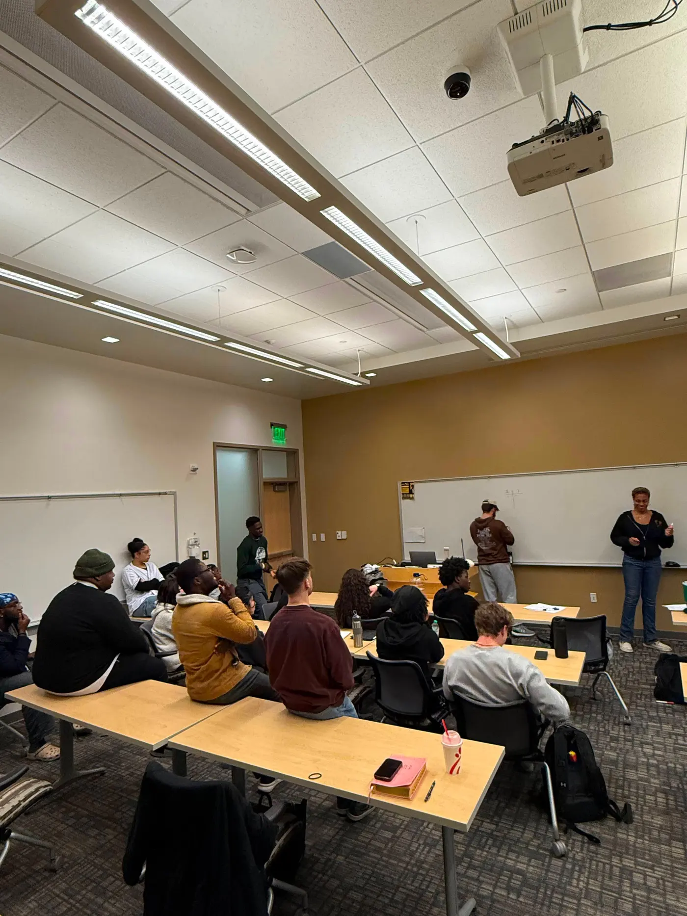 Students seated at long classroom tables, facing two people standing at the front of the room.