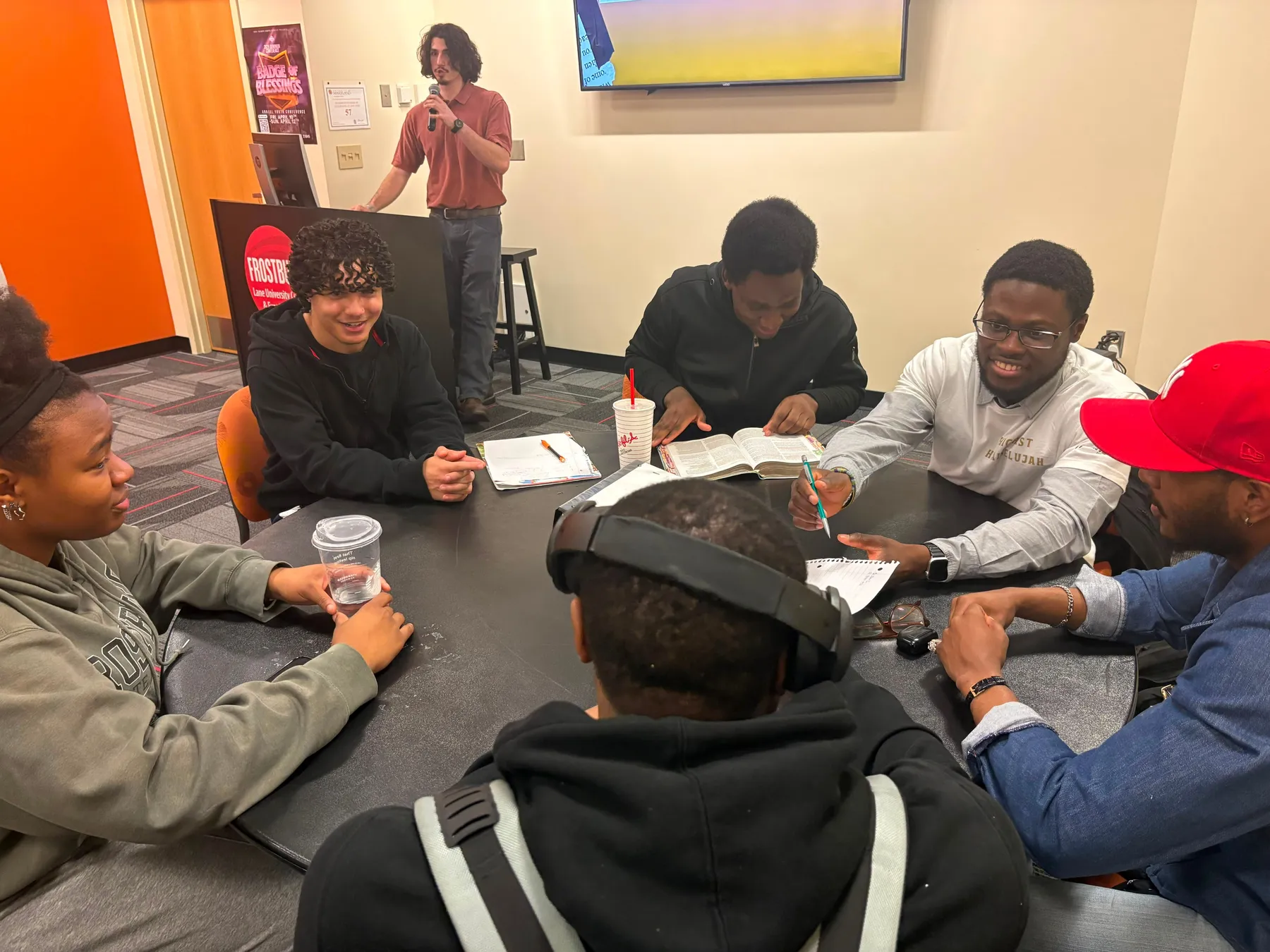 Students seated around a table with open Bibles and workbooks while one person speaks at the front of the room.