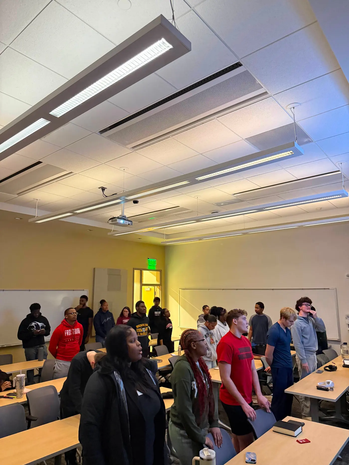 A classroom of students on their feet facing the front of the room under long strip lights.