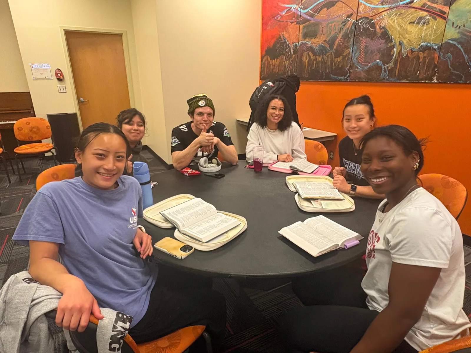 Six students seated around a round table with open Bibles in front of them, looking up at the camera.