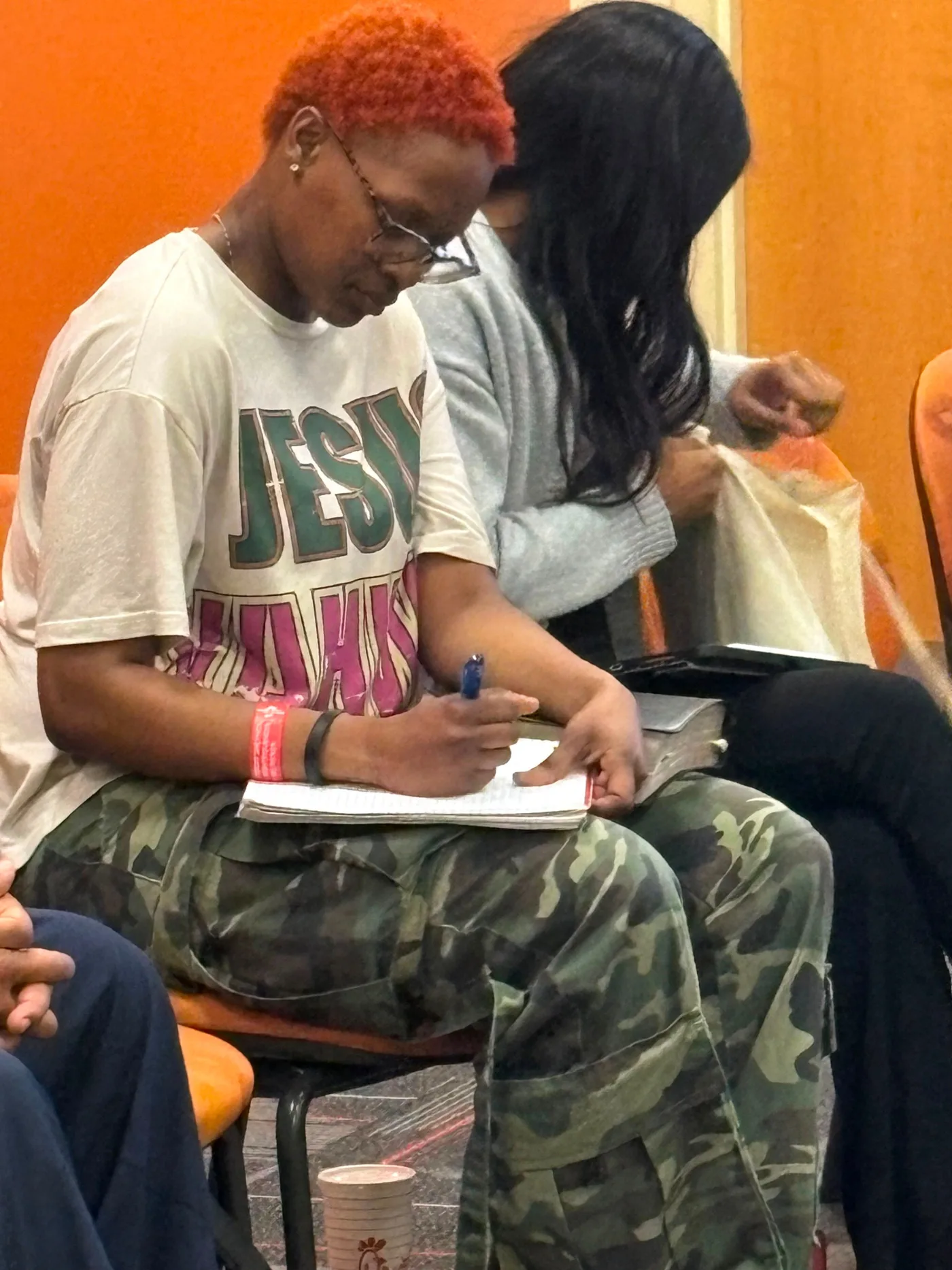 A student writing in an open notebook balanced on their lap, seated among others in a bright room.