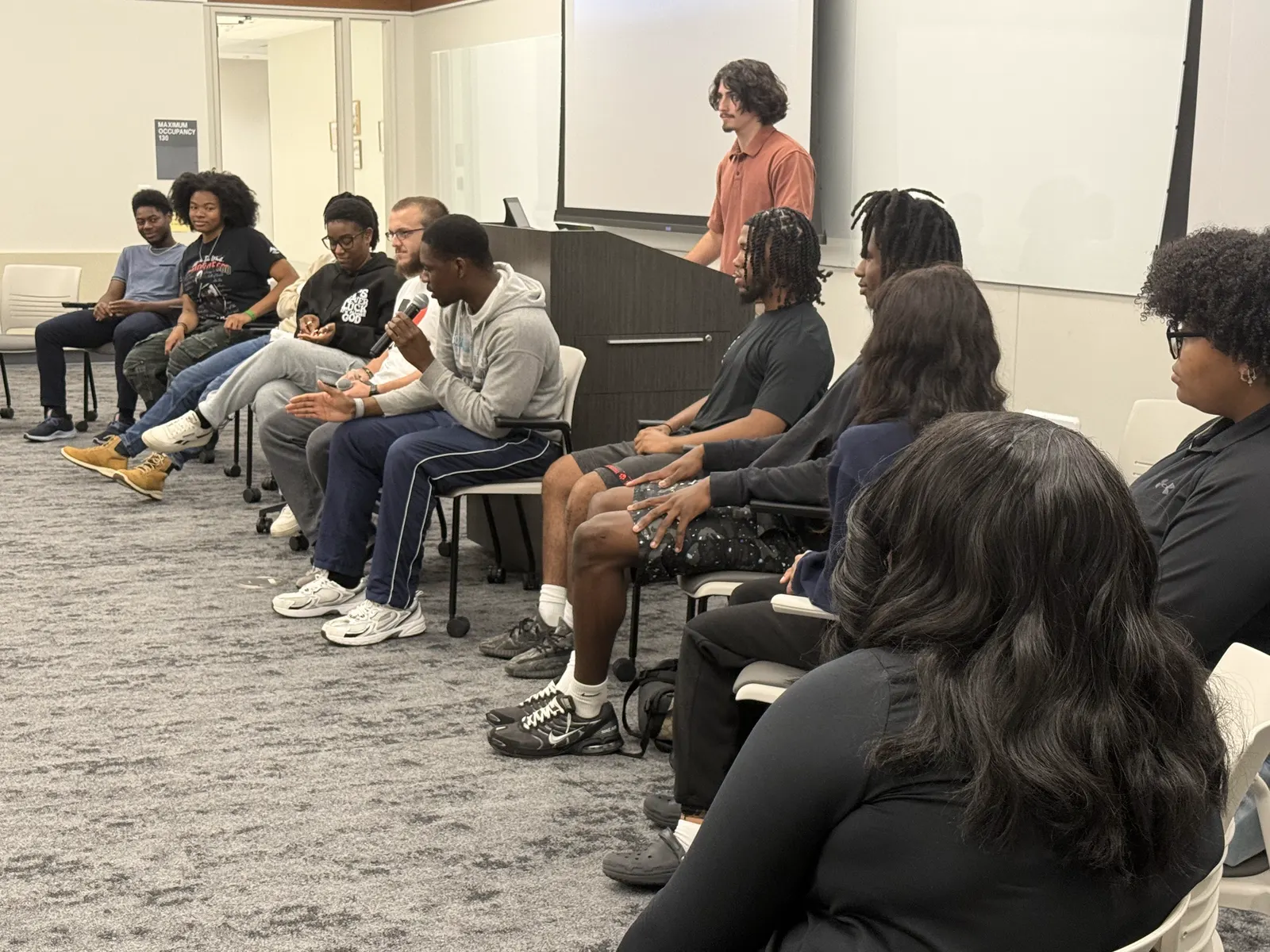Students seated in a row along a wall, listening to a person standing at the front of the room.