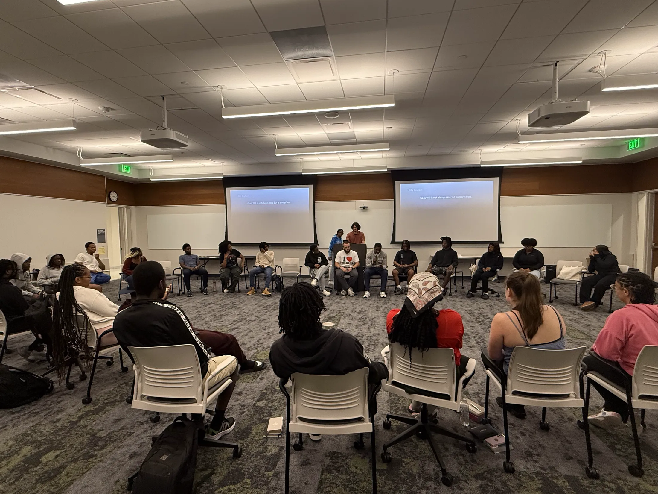 Students seated in a wide circle of chairs filling a meeting room, with two lit screens at the front.