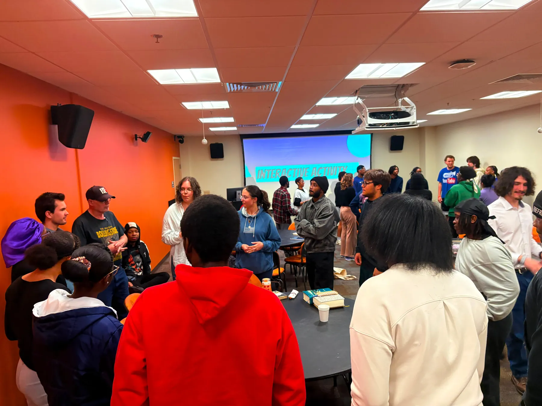 A meeting room full of students standing and talking in groups, with a lit screen at the far end.
