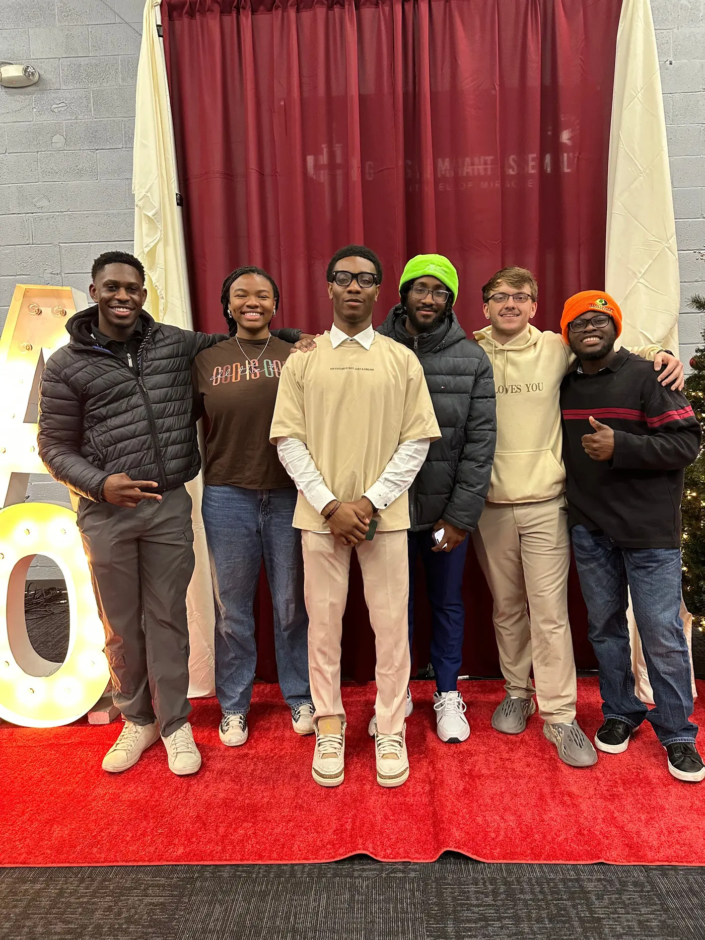 Six students standing on a red carpet in front of a curtain, beside a large lit letter.