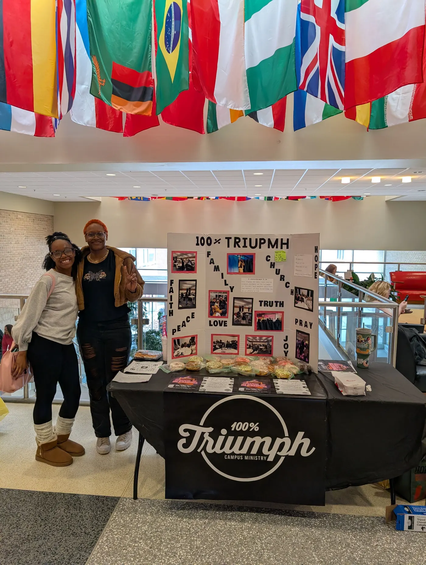 Two students standing beside a 100% Triumph table on campus, a display board behind them and international flags hanging overhead.