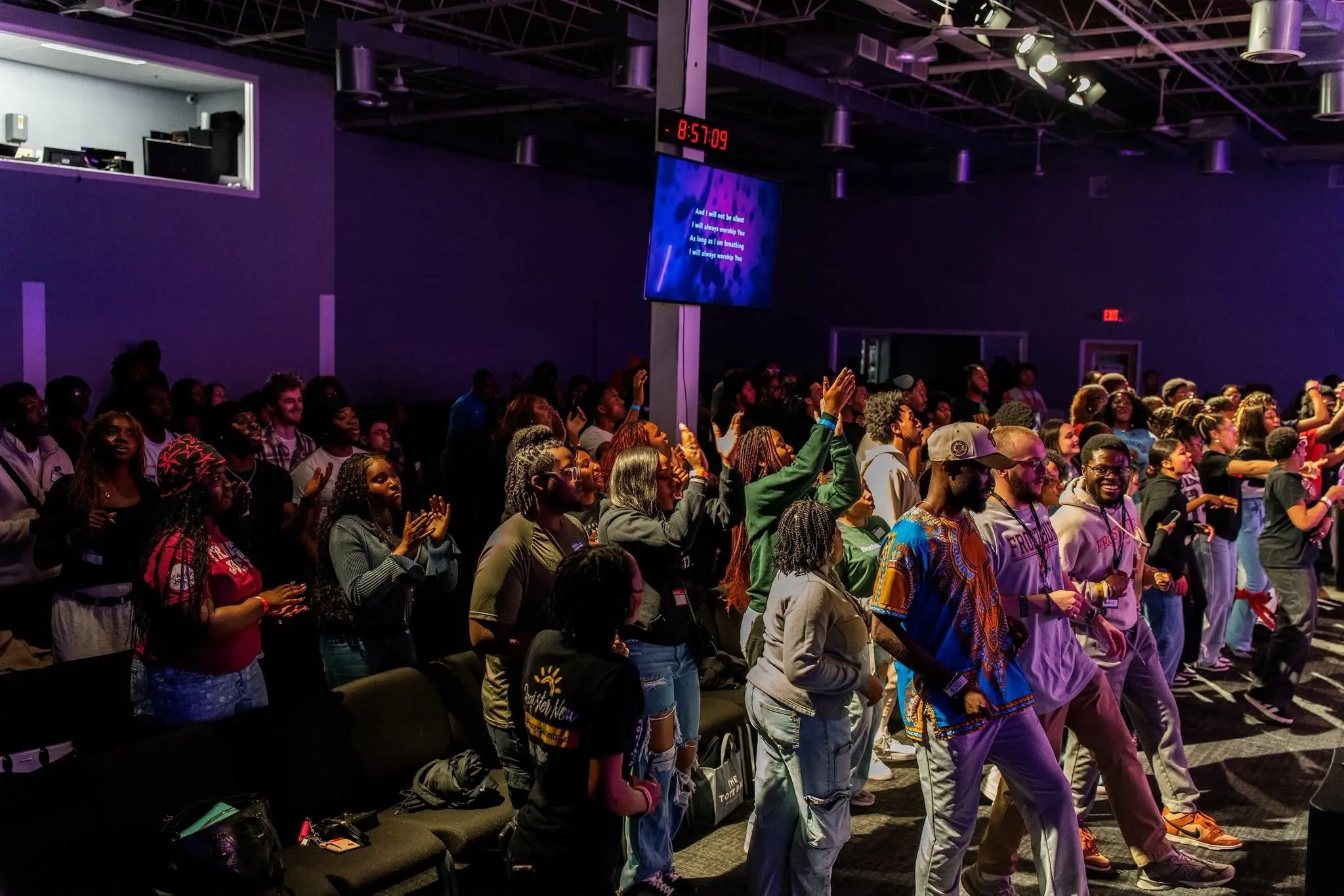 Students filling a dark hall lit purple, standing close together with several arms raised.
