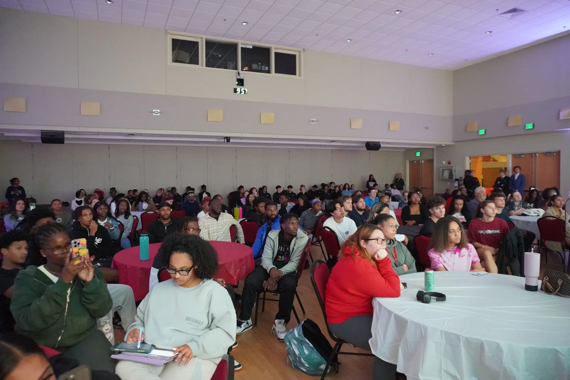 Rows of students seated at round tables in a bright hall, watching the front of the room.
