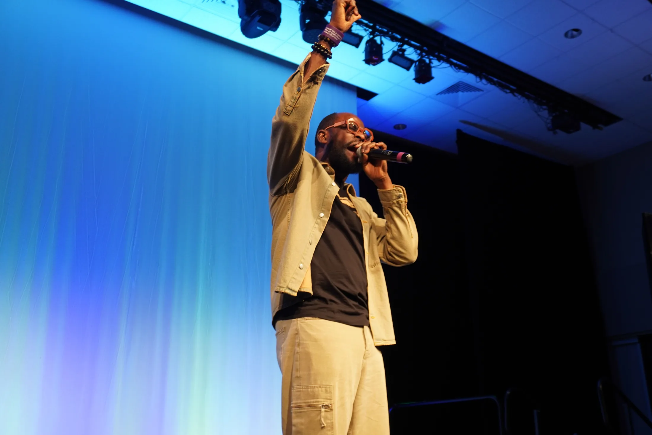 A speaker on a lit stage holding a microphone with one arm raised, against a backdrop washed blue and green by the stage lighting.
