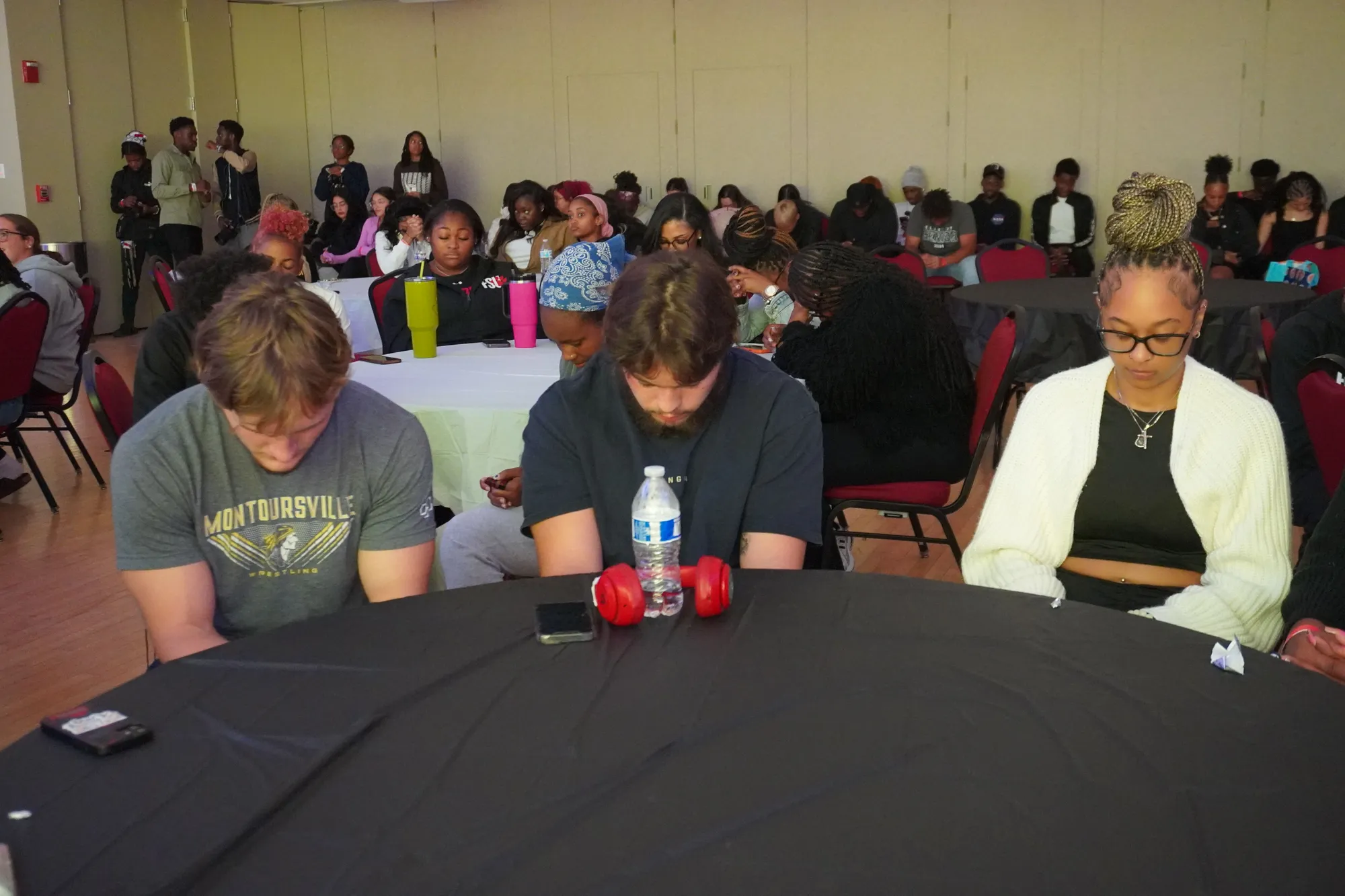 Students at the front tables of a full hall with their heads bowed over the table in front of them.