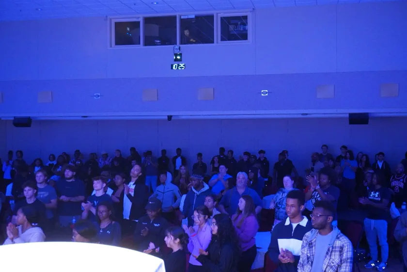 A hall of students on their feet in deep blue light, facing the front of the room, many with hands raised or clasped in front of them.