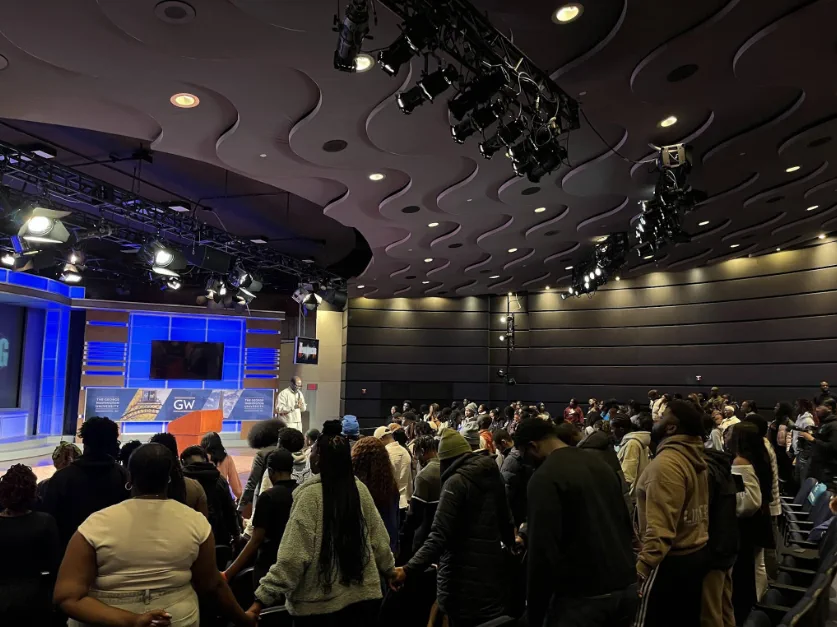 Students standing shoulder to shoulder at the back of the auditorium, many with heads bowed and hands joined, while the speaker stands at the lectern on the lit stage beyond them.