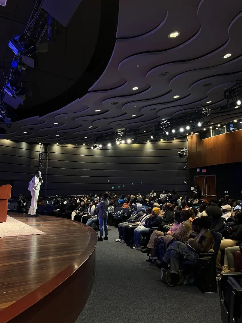 A speaker stands at the curved edge of the stage with a microphone while every seat in the raked auditorium in front of them is taken, under a sculpted ceiling hung with stage lighting.