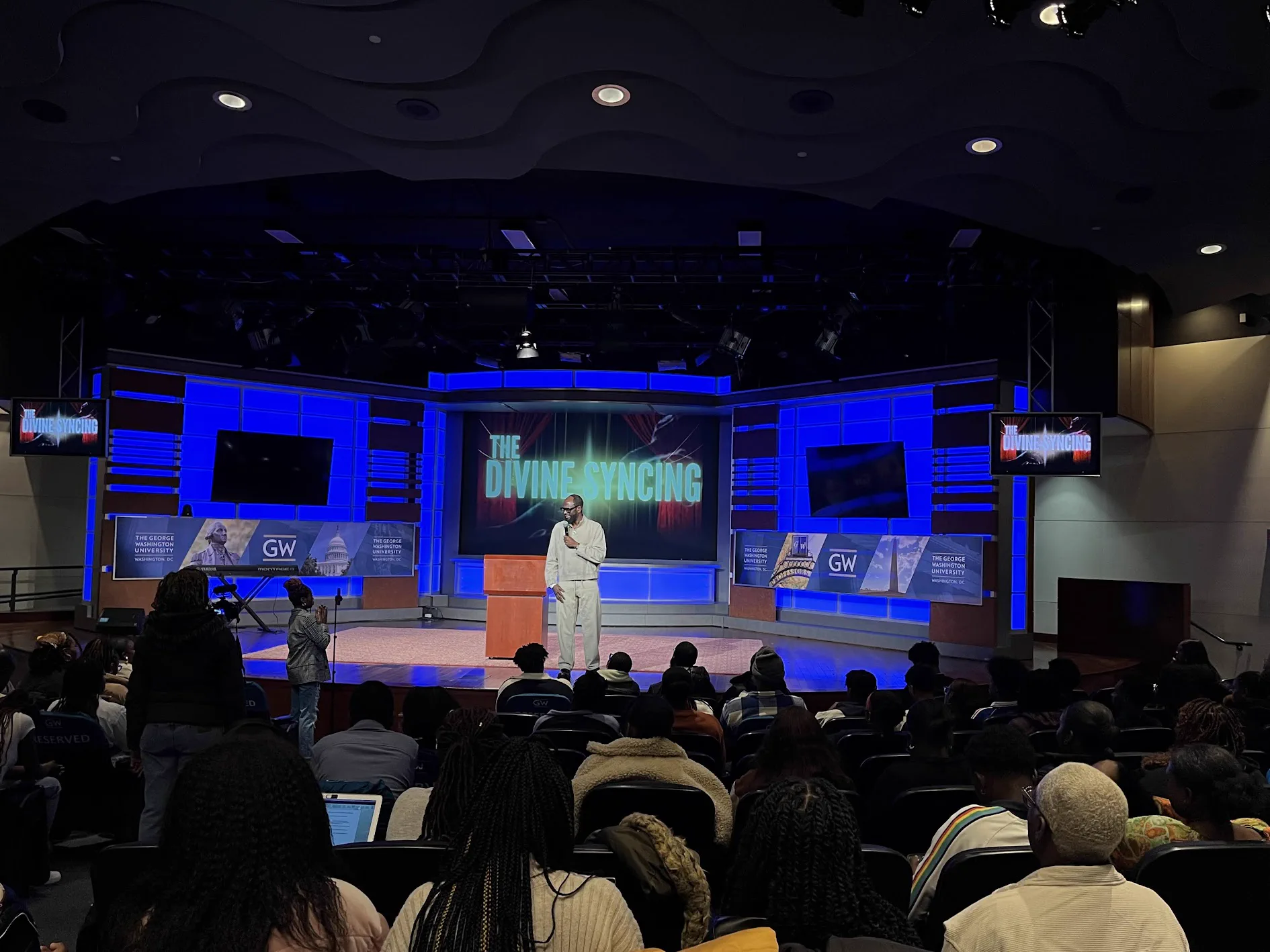 A Dunamis Conference session seen from the back of a full auditorium. A speaker in a white suit stands at the front of a blue-lit stage holding a microphone, beside a screen reading "The Divine Syncing", with George Washington University banners on either side.