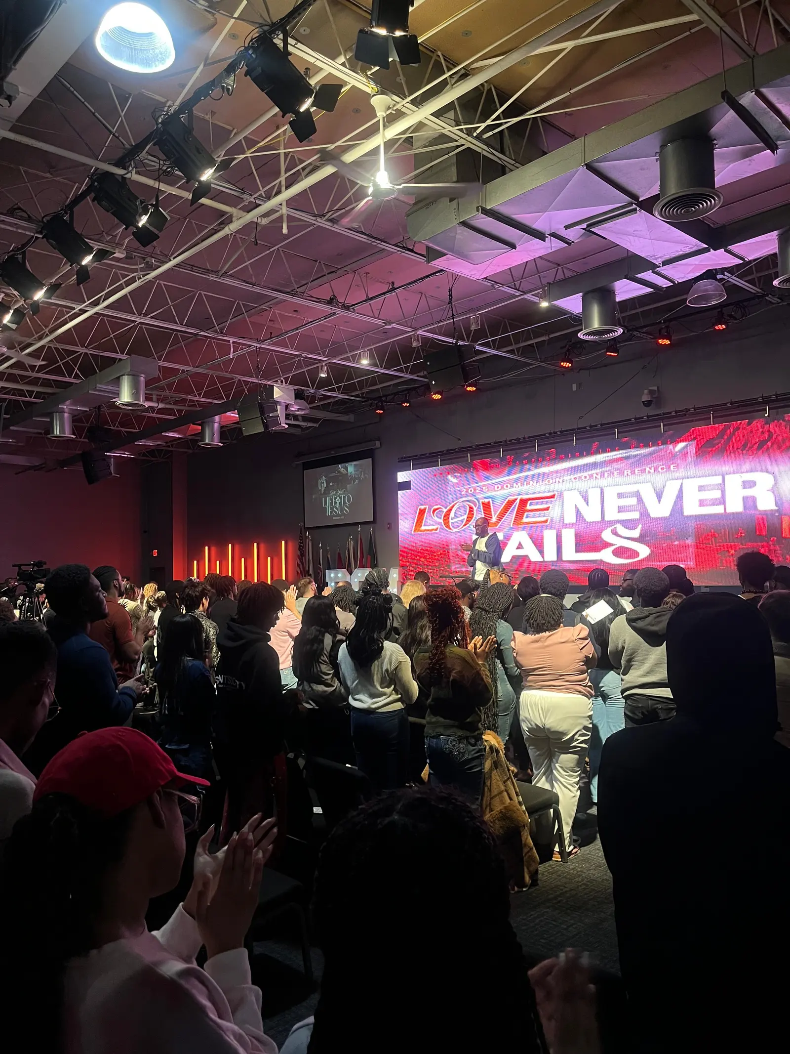 A conference hall seen from the back rows, the crowd on their feet under pink and red stage lighting, with "Love Never Fails" lit on the screen at the front.