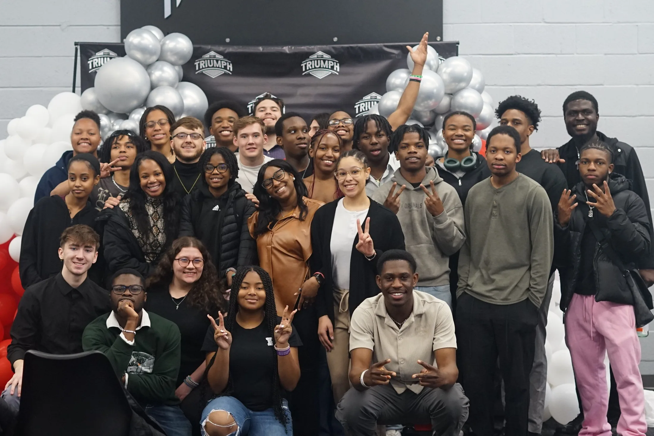 A large group of students crowded together in front of a 100% Triumph backdrop hung with balloons, smiling at the camera.